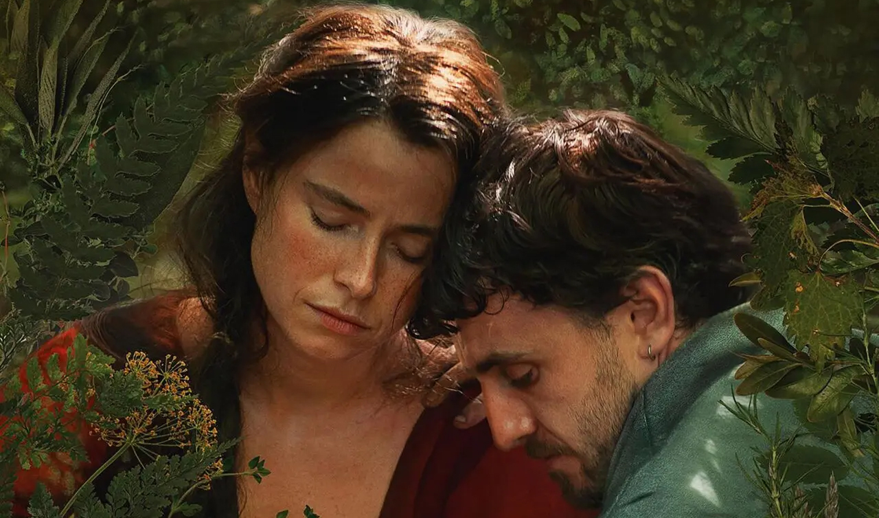 A woman and a man sit close together among green foliage, their heads gently leaning toward each other, eyes closed, conveying a quiet, emotional moment in natural light.