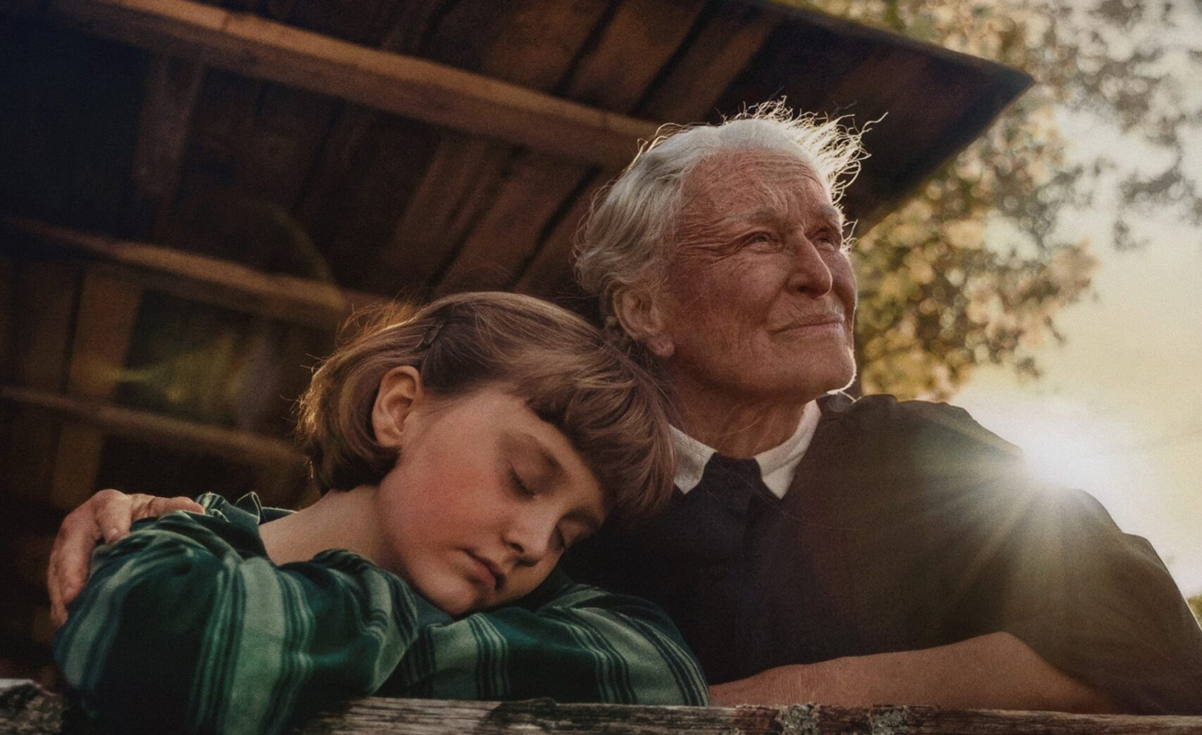 A young girl and an old woman are leaning on the outer wall of a wooden porch. The girl is asleep, her head resting on her arm. It is late summer and the afternoon sun shines behind them.